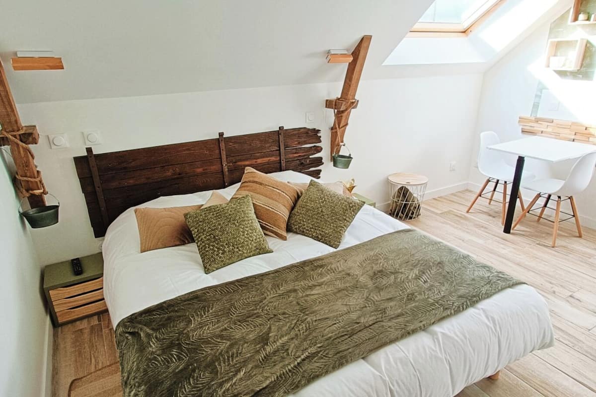 A bedroom with a wooden headboard, a large bed with multiple pillows, a bedside table, a small round table, and a white chair. There is a skylight above the bed.