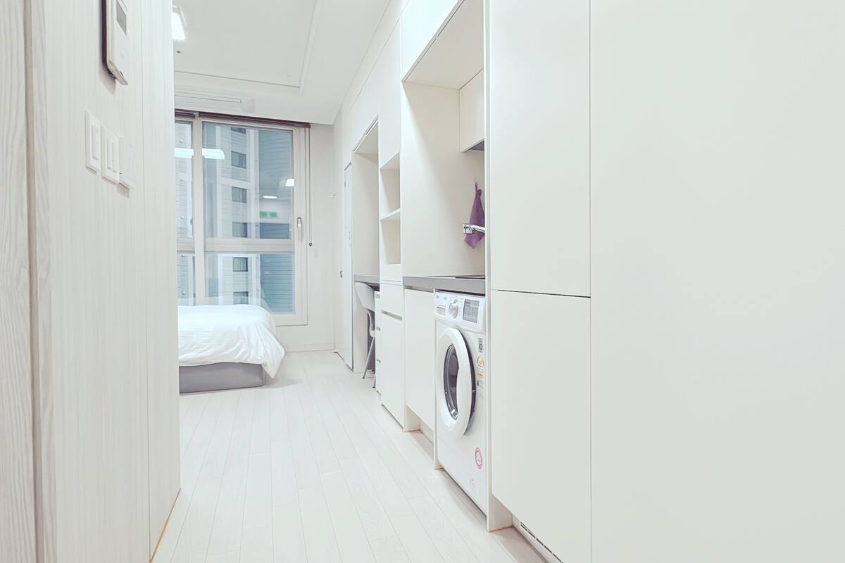 A modern, white-walled bedroom with a large window, a washing machine, and a bed.
