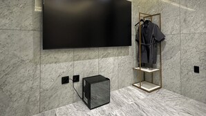 A modern bathroom with a large flat-screen TV mounted on the wall, a small black refrigerator, and a robe rack with clothes. - Hotel Island (Ansan)