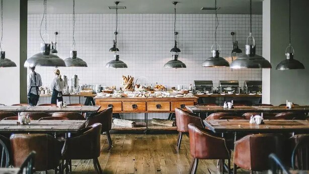 Un restaurante espacioso con mesas y sillas de madera, una isla central con equipo de cocina, y lámparas colgantes de estilo industrial.