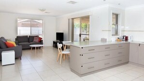 Una cocina moderna con una isla central, un comedor con mesa y sillas blancas, y una sala de estar con sofá y televisor.