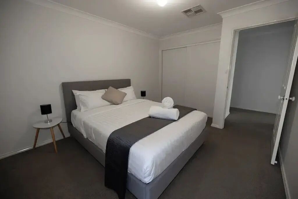 Una cama bien tendida con cabecera gris y ropa de cama blanca, una mesita de noche con lámpara y una puerta cerrada.