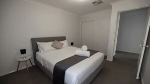 Una cama bien tendida con cabecera gris y ropa de cama blanca, una mesita de noche con lámpara y una puerta cerrada.