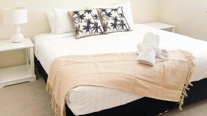 Una cama bien hecha con edredón blanco, una manta color beige y un par de toallas dobladas al lado.