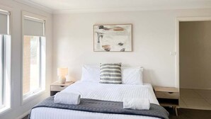 A bedroom with a bed, a nightstand, a lamp, a painting on the wall, and a window. - Welcome to our stylish 3BR villa, perfect for an escape (Wagga Wagga)
