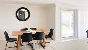 A modern dining area with a wooden table and black chairs, a round mirror on the wall, and a sliding glass door leading to an outdoor area. - Welcome to our stylish 3BR villa, perfect for an escape (Wagga Wagga)