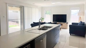 A modern kitchen with a dishwasher, a sink, and a built-in TV. There is a dark-colored sofa and a lamp in the living area. - Welcome to our stylish 3BR villa, perfect for an escape (Wagga Wagga)
