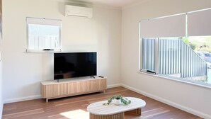 Een moderne woonkamer met een flatscreen televisie op een houten kast, een kleine ronde tafel met een plant en een groot raam met uitzicht.