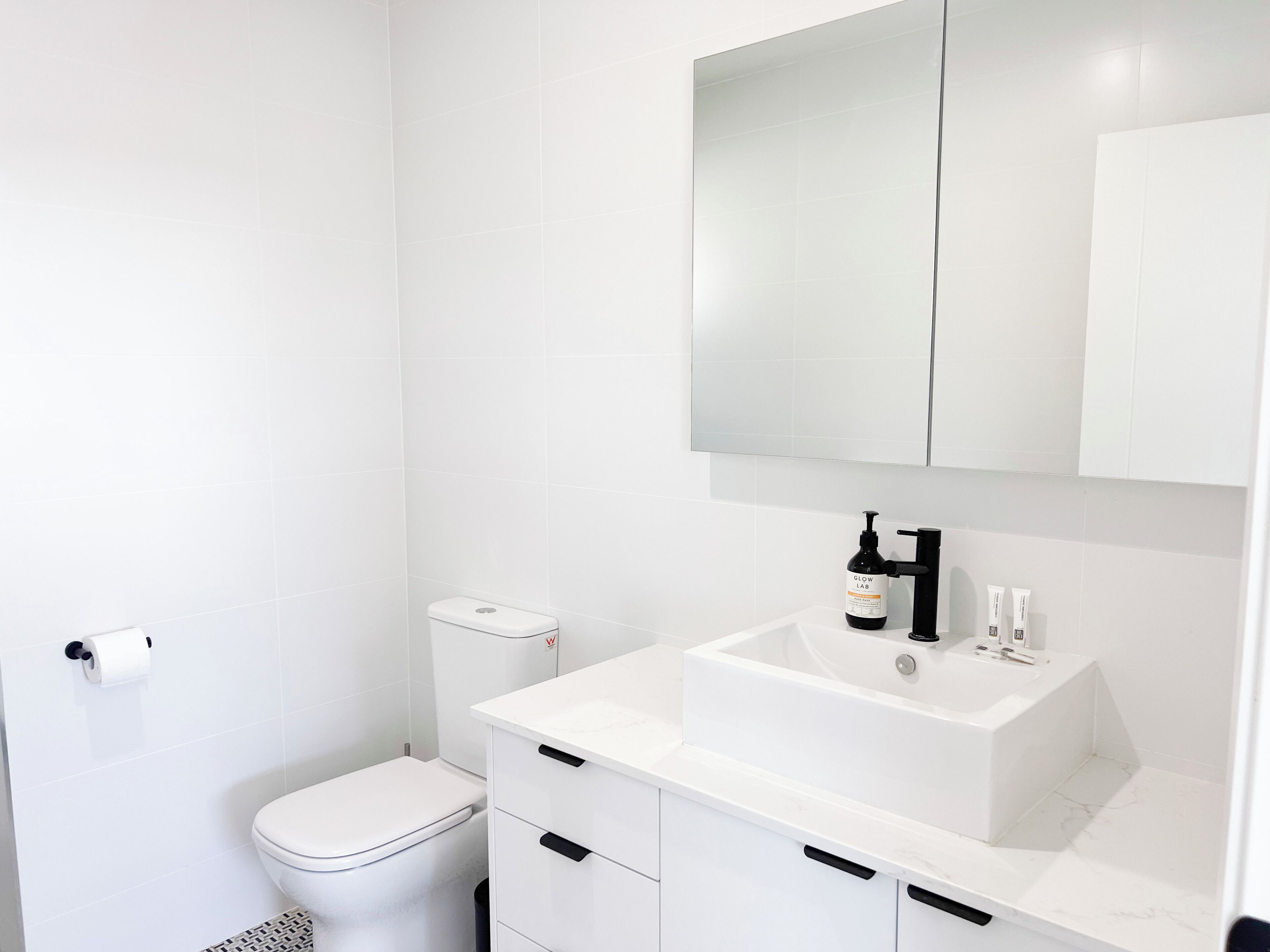 Une salle de bain moderne avec des toilettes, un lavabo et un miroir au-dessus, ainsi qu’une bouteille de shampoing.