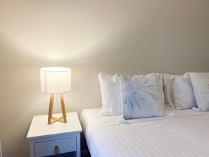 Ein ordentlich bezogenes Bett mit weißer Bettwäsche und einem dekorativen Kissen mit Blattmuster. Ein weißer Nachttisch mit Lampe und Schublade steht daneben.