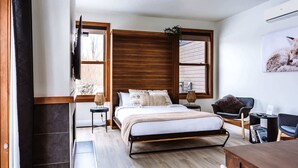 A modern bedroom with a large bed, wooden headboard, two bedside tables, a chair, a desk, and a window with a view.