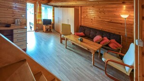 A wooden cabin interior with a dining table, chairs, a sofa with red cushions, a television, and a lamp.