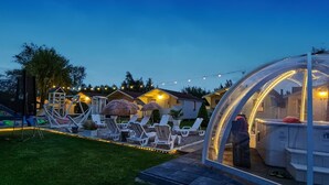 A cozy outdoor seating area with lounge chairs and a thatched-roof structure, surrounded by houses with string lights.