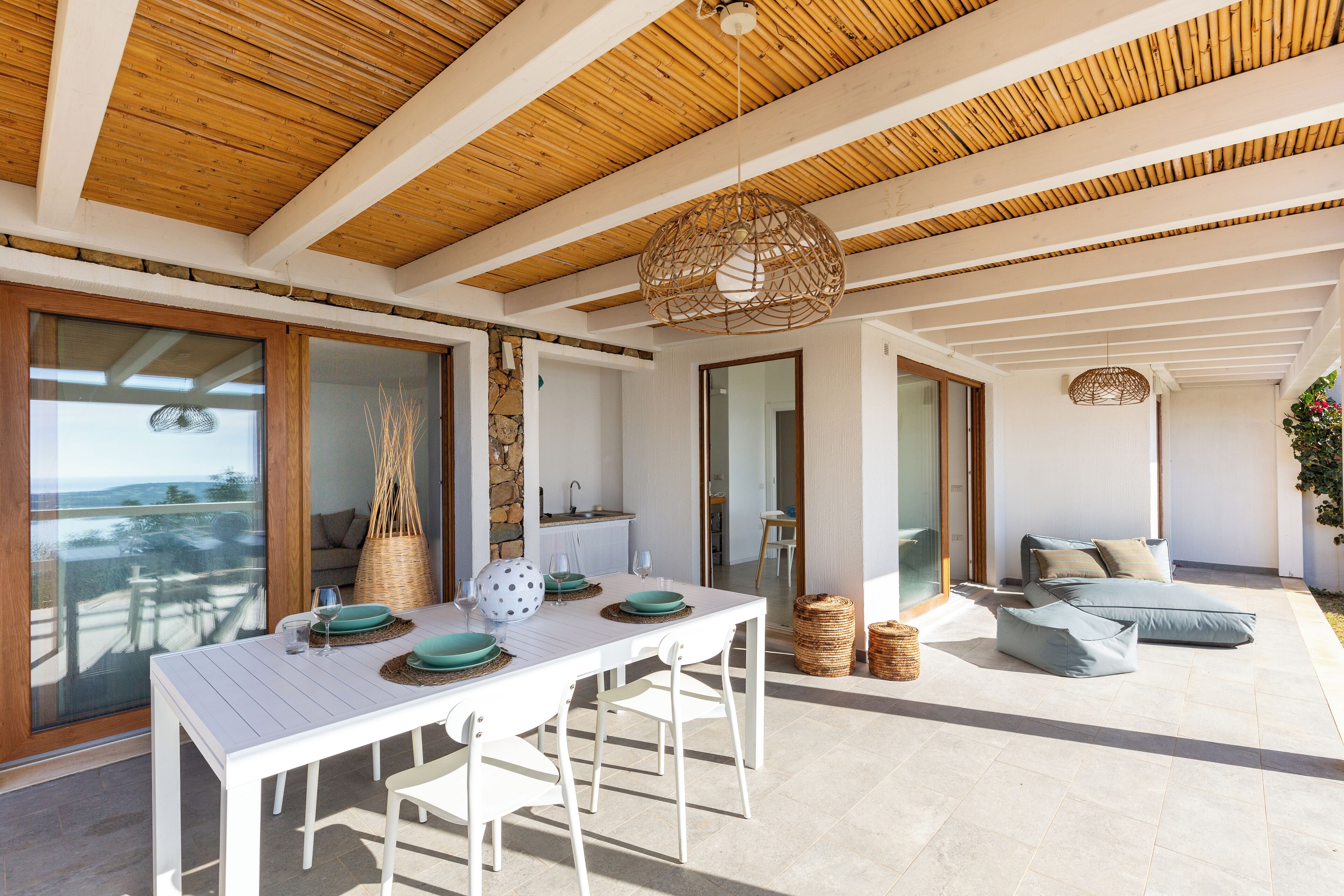 A modern outdoor dining area with a white table and chairs, a wicker hanging light fixture, and a view of the sea.