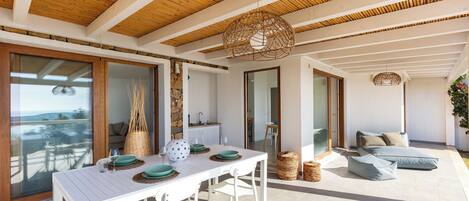 A modern outdoor dining area with a white table and chairs, a wicker hanging light fixture, and a view of the sea.