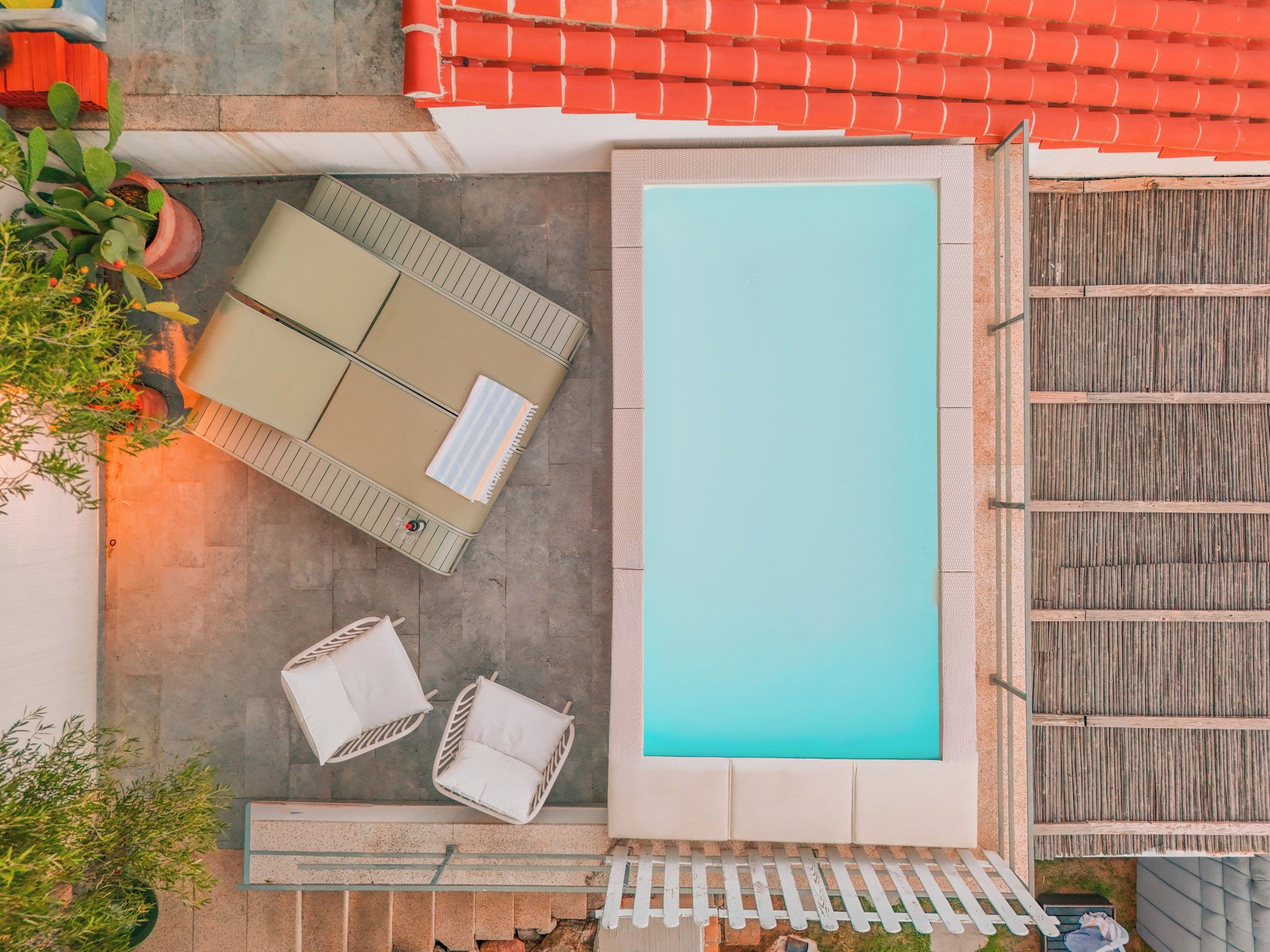 Ein Poolbereich auf dem Dach mit einem rechteckigen Pool, zwei weißen Liegestühlen, einem kleinen Tisch mit Kissen und Blumentöpfen.