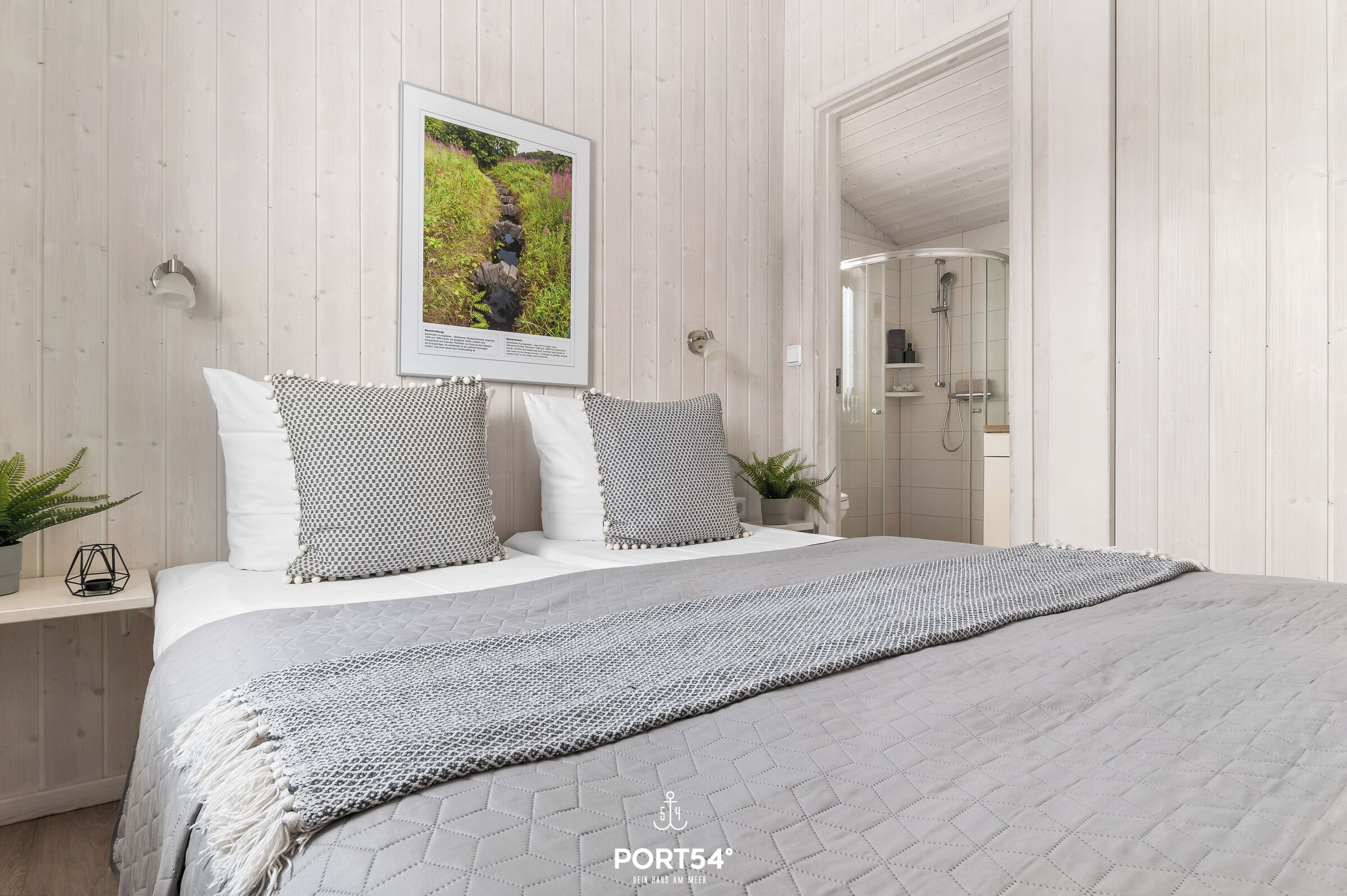 Une chambre à coucher avec un lit, des oreillers, un tableau encadré, une plante sur la table de chevet et un espace douche.