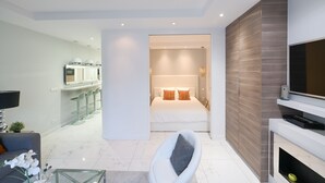 Habitación de hotel moderna con cama, televisión, zona de estar y cocina visible a través de una puerta abierta.