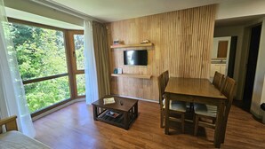 A modern living room with a wooden dining table and chairs, a flat-screen TV mounted on a wooden panel wall, and a large window offering a view of greenery.