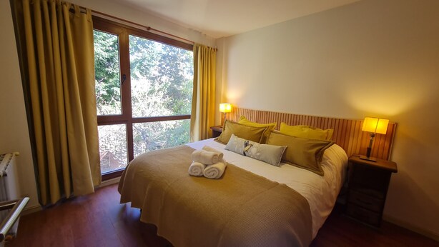 A hotel room with a large window, a bed with a beige bedspread, multiple pillows, and two bedside lamps.