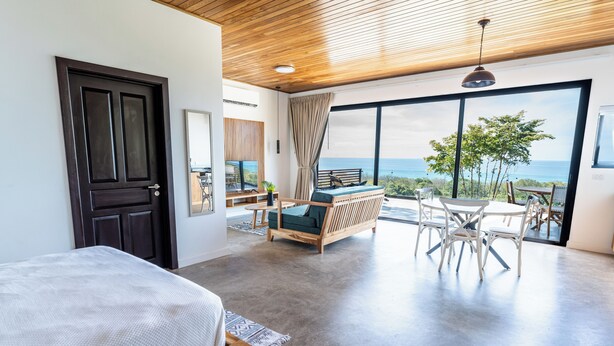 Une chambre à coucher moderne avec un grand lit, un plafond en bois et une vue sur l’océan grâce à de grandes fenêtres.
