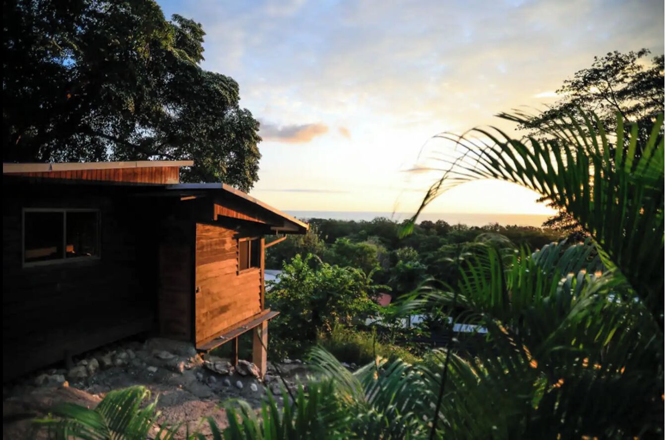 A wooden cabin with a sloped roof, surrounded by lush greenery and a view of the sunset.