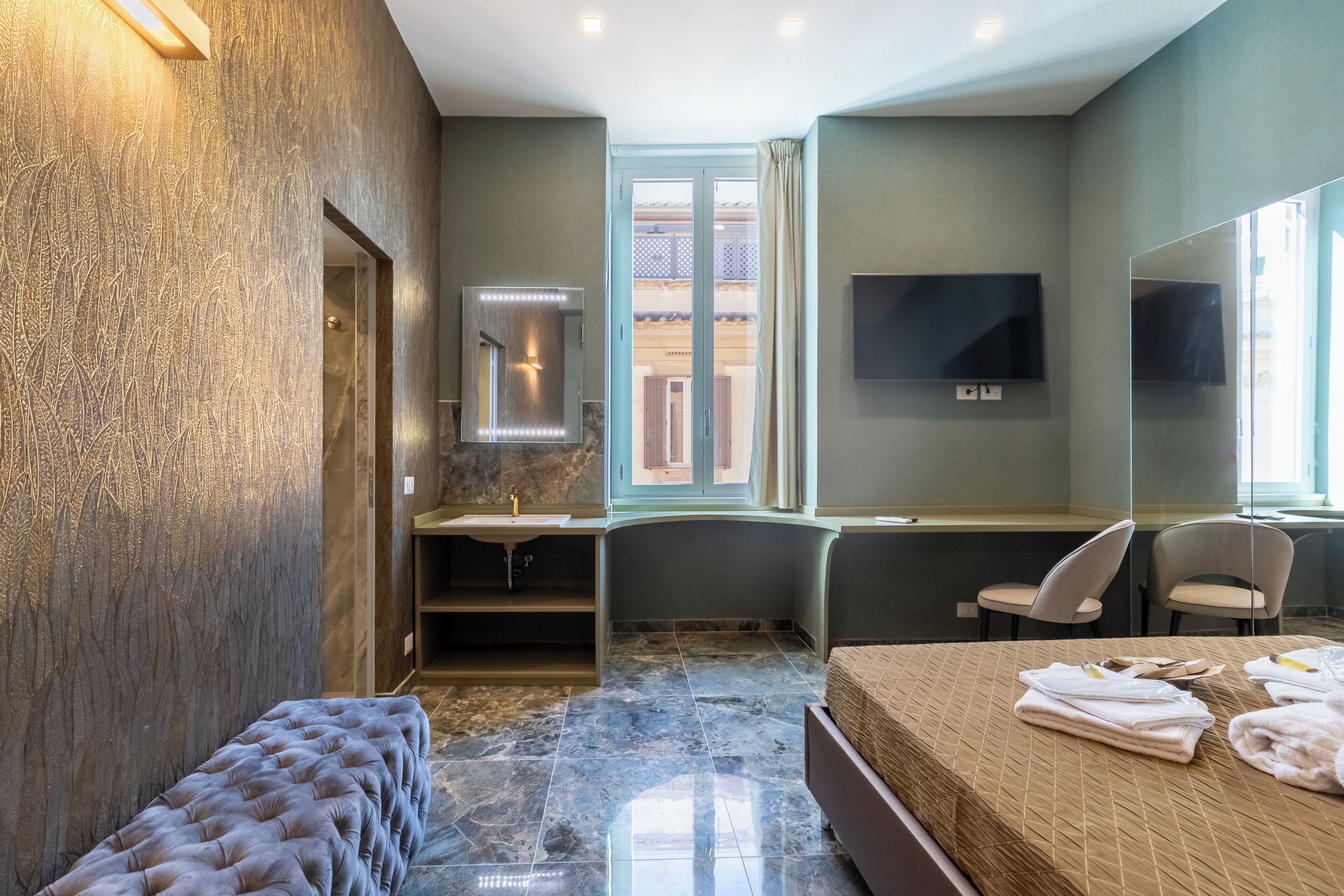 A modern hotel room with a large bed, a vanity with a mirror, a bathtub, and a seating area with chairs.
