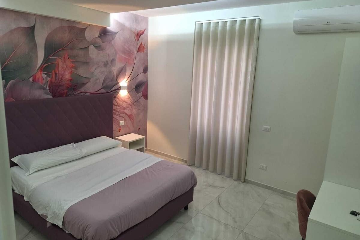 A bedroom with a bed, a nightstand, a wall-mounted air conditioner, and a window with curtains.