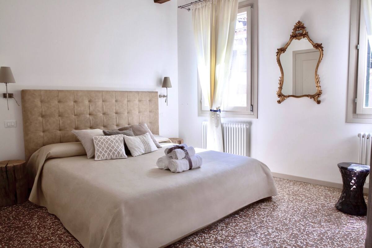 A neatly made bed with a tufted headboard, a mirror, and a window with curtains.