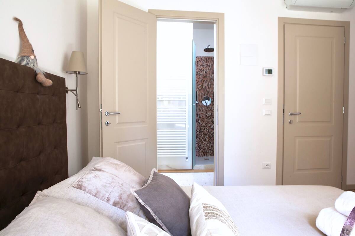 A bedroom with a tufted headboard, a bed with multiple pillows, a bedside lamp, and a door leading to a bathroom with a shower and a tiled wall.
