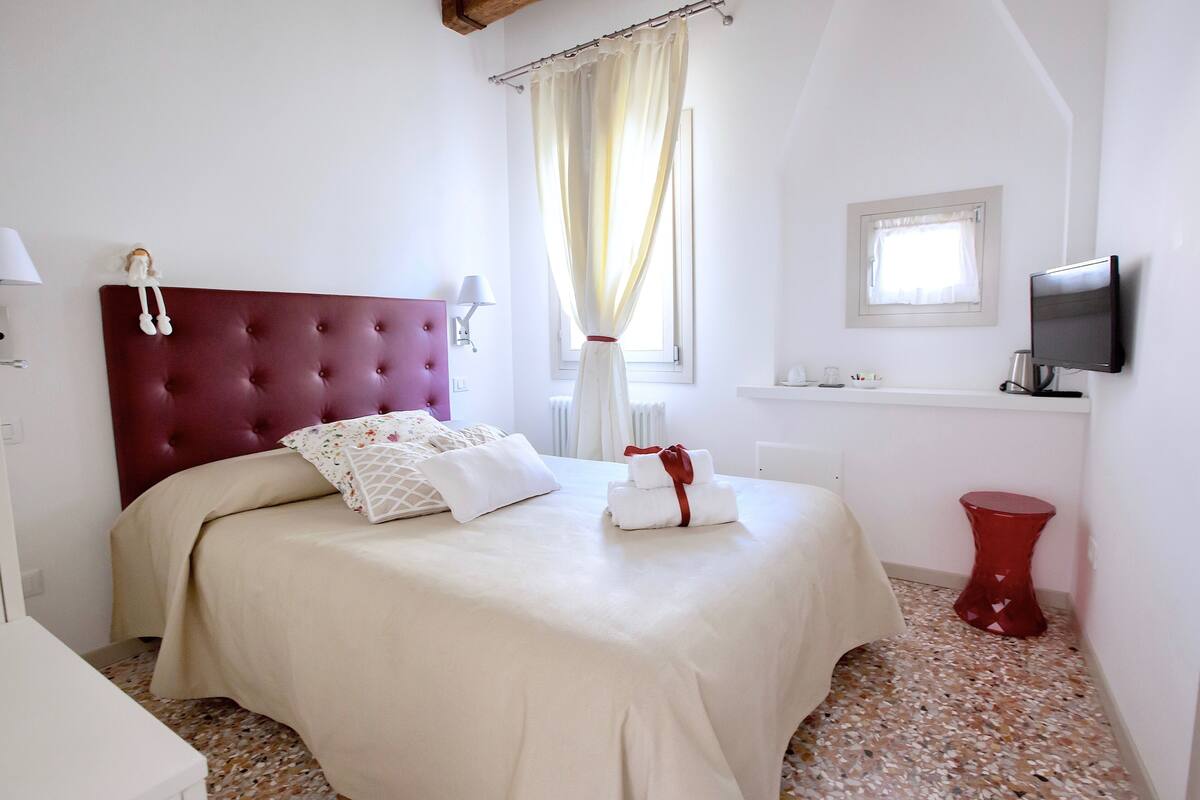 A neatly made bed with a red headboard, a bedside table with a lamp, a television mounted on the wall, and a window with curtains.
