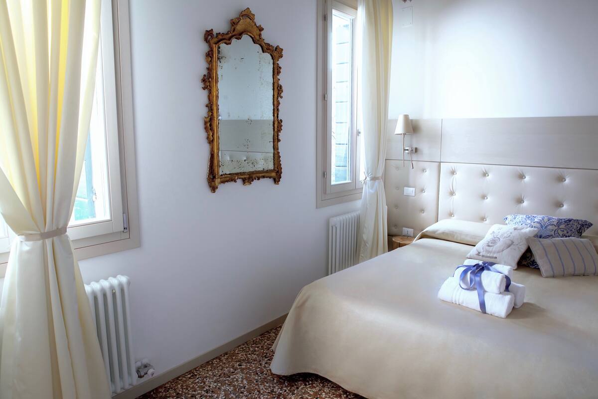 A hotel room with a large bed, a window with curtains, a radiator, and a wall-mounted mirror.