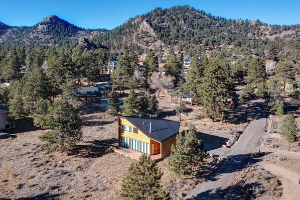 Una casa moderna con techo inclinado, rodeada de un área boscosa y una montaña al fondo.