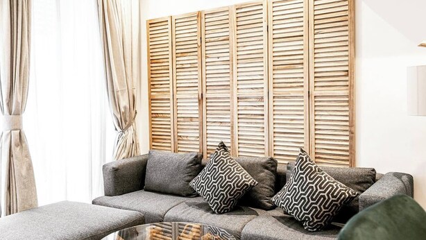 Ein modernes Wohnzimmer mit einem grauen Sofa, gemusterten Kissen, einem Glastisch mit Holzfuß und einem gewebten Teppich.