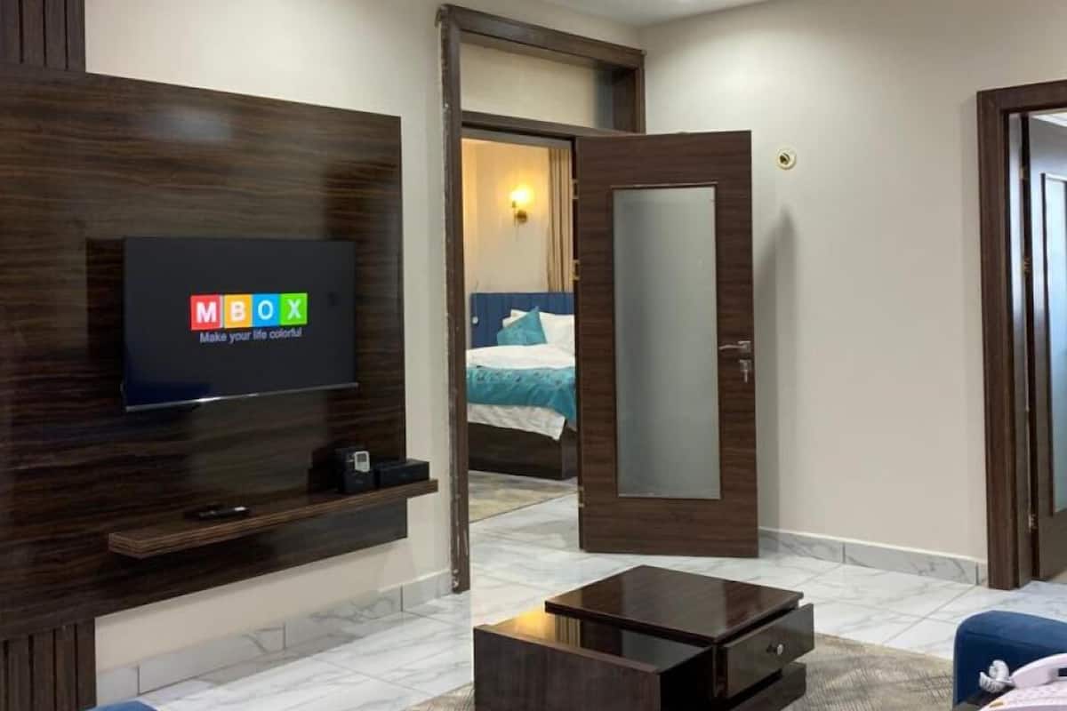 A modern hotel room with a large flat-screen TV mounted on a wooden wall, a bed with blue bedding, a small bedside table, and a mirror above the bed.