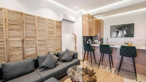 Ein modernes Wohnzimmer mit grauem Sofa, gemusterten Kissen und einem Glastisch. Es gibt eine Küchenzeile mit Bar und Hockern sowie einen großen Spiegel an der Wand.