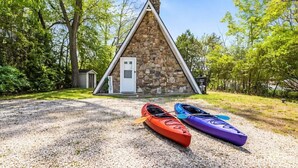 A stone cabin with a triangular roof, two kayaks in front, surrounded by trees.