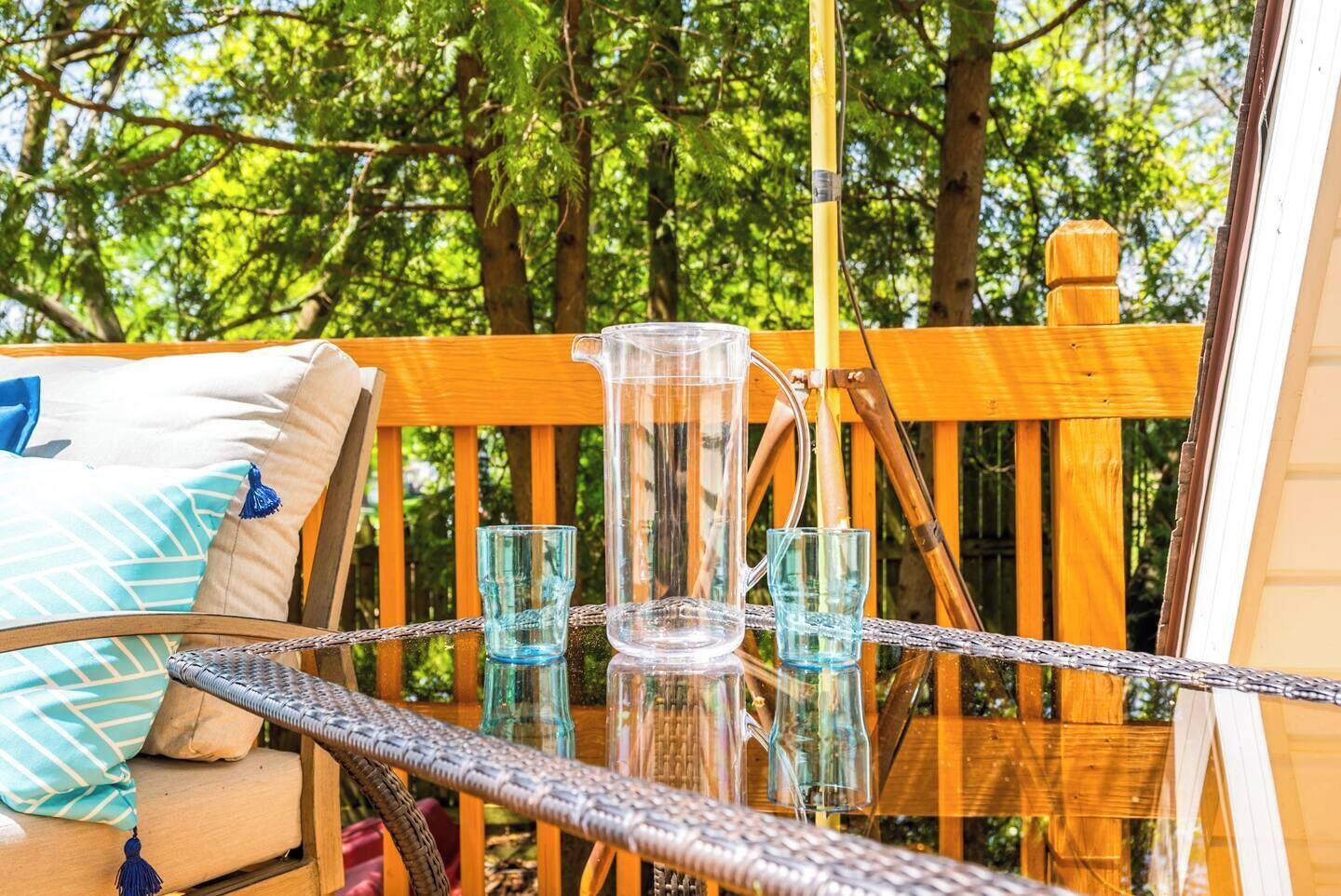 A glass table with a pitcher and two glasses, set on a wooden deck with a view of trees.