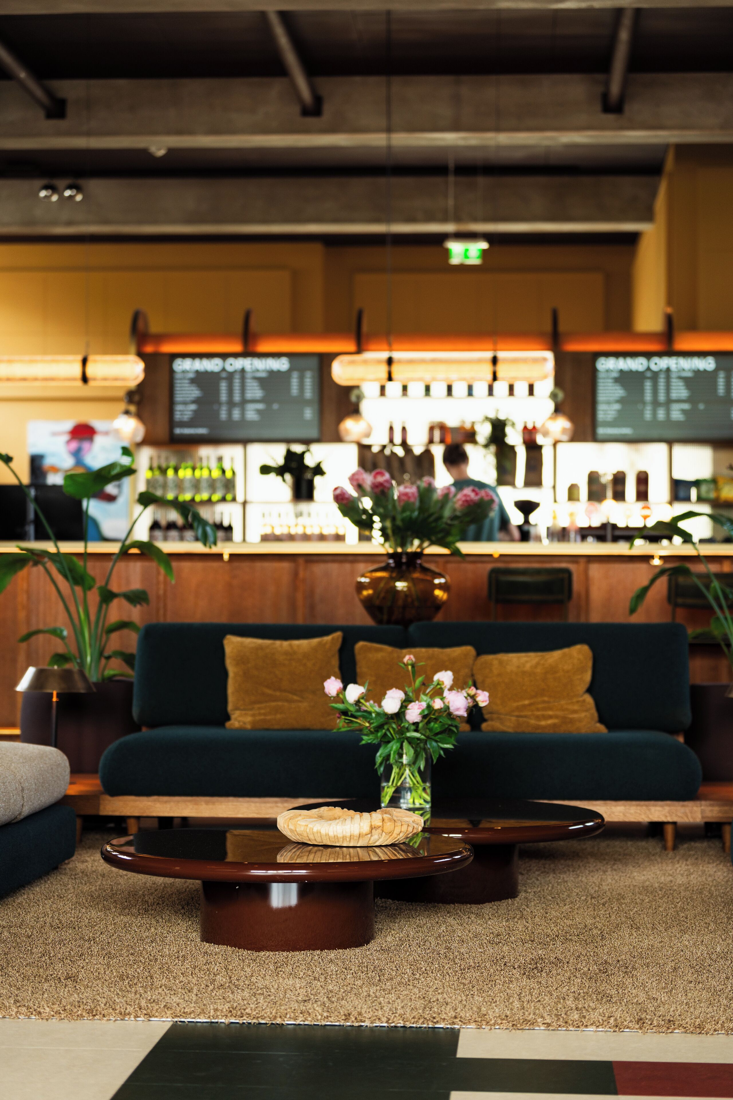 A bar area with a dark green sofa, a wooden bar counter, and a vase with flowers on a coffee table.