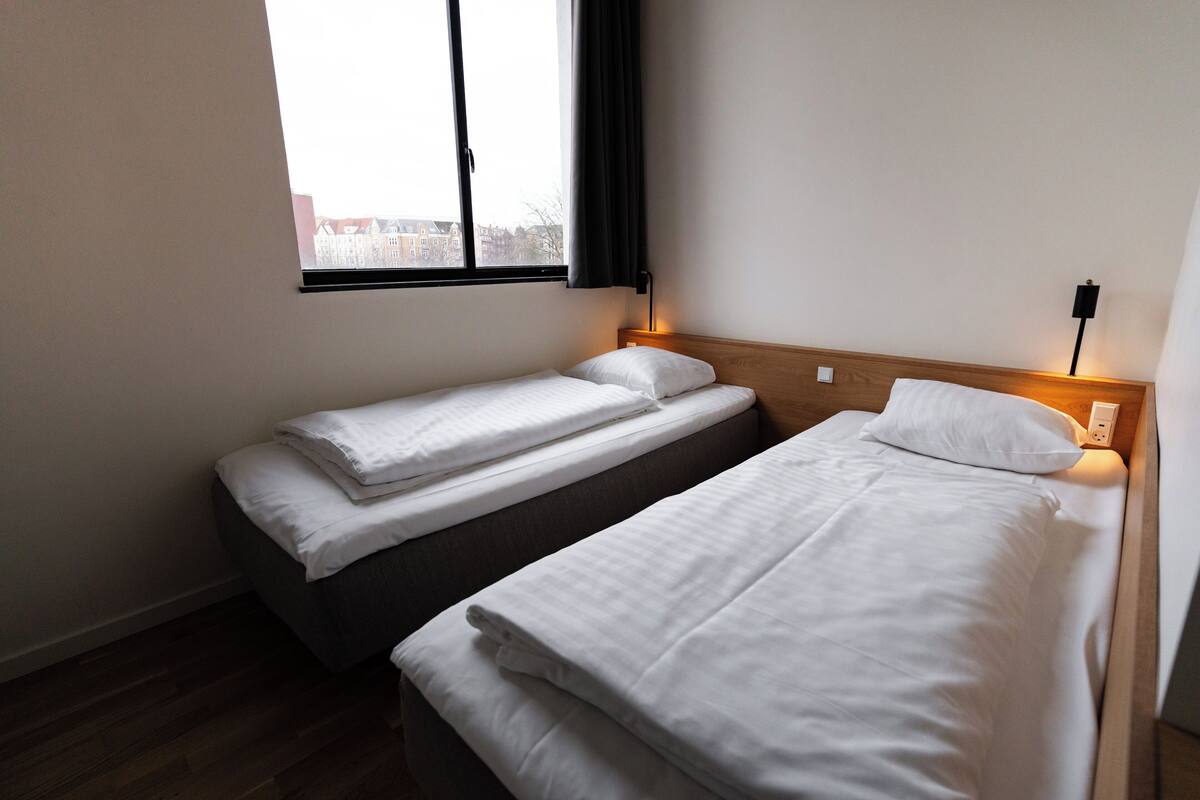 A small, clean room with two single beds, a window with a view of buildings, and a bedside table with a lamp.