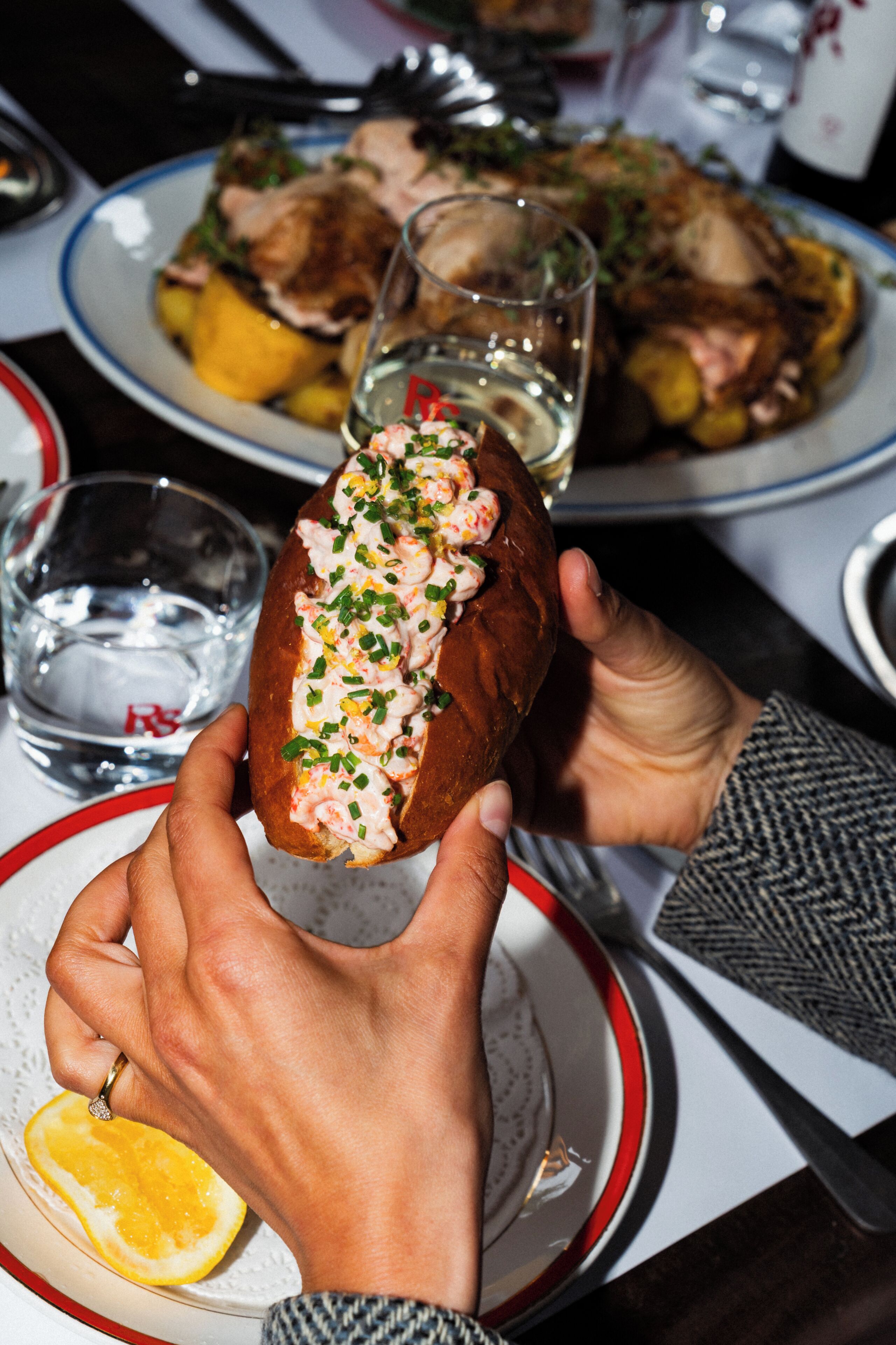 En person holder en lobster roll med et glas vand og en citronskive på et bord.