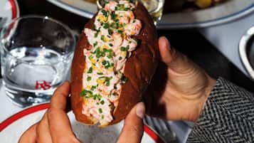 En person holder en lobster roll med et glas vand og en citronskive på et bord.