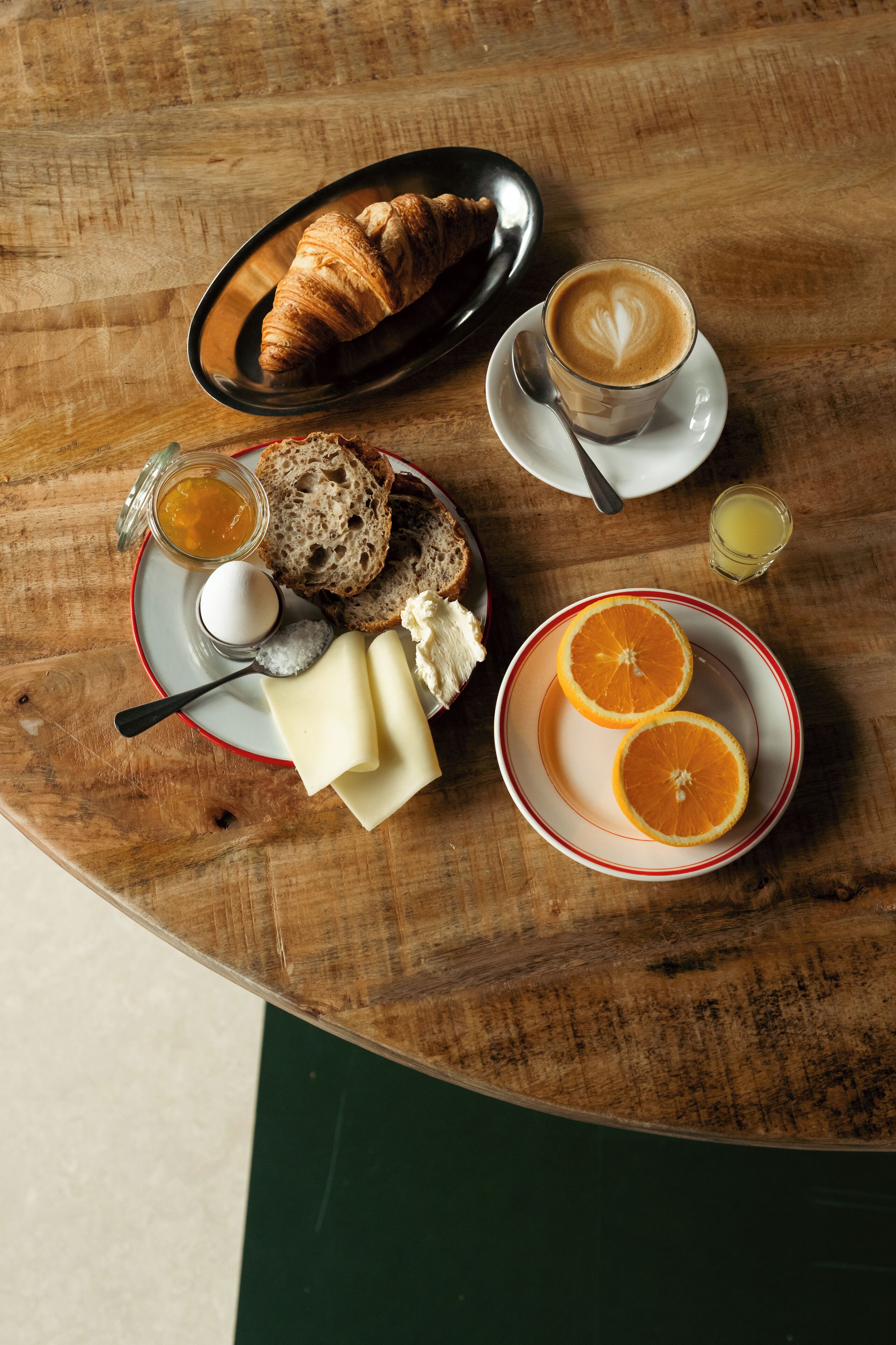 A wooden table with a croissant, a glass of coffee, a plate with cheese and bread, and a plate with orange slices.