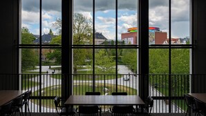 A room with a large window overlooking a courtyard with trees and a building. There is a table and chairs in the foreground. - Robertas Society (Aarhus)