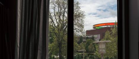 A window with curtains, a view of a park, and a building with a green roof.