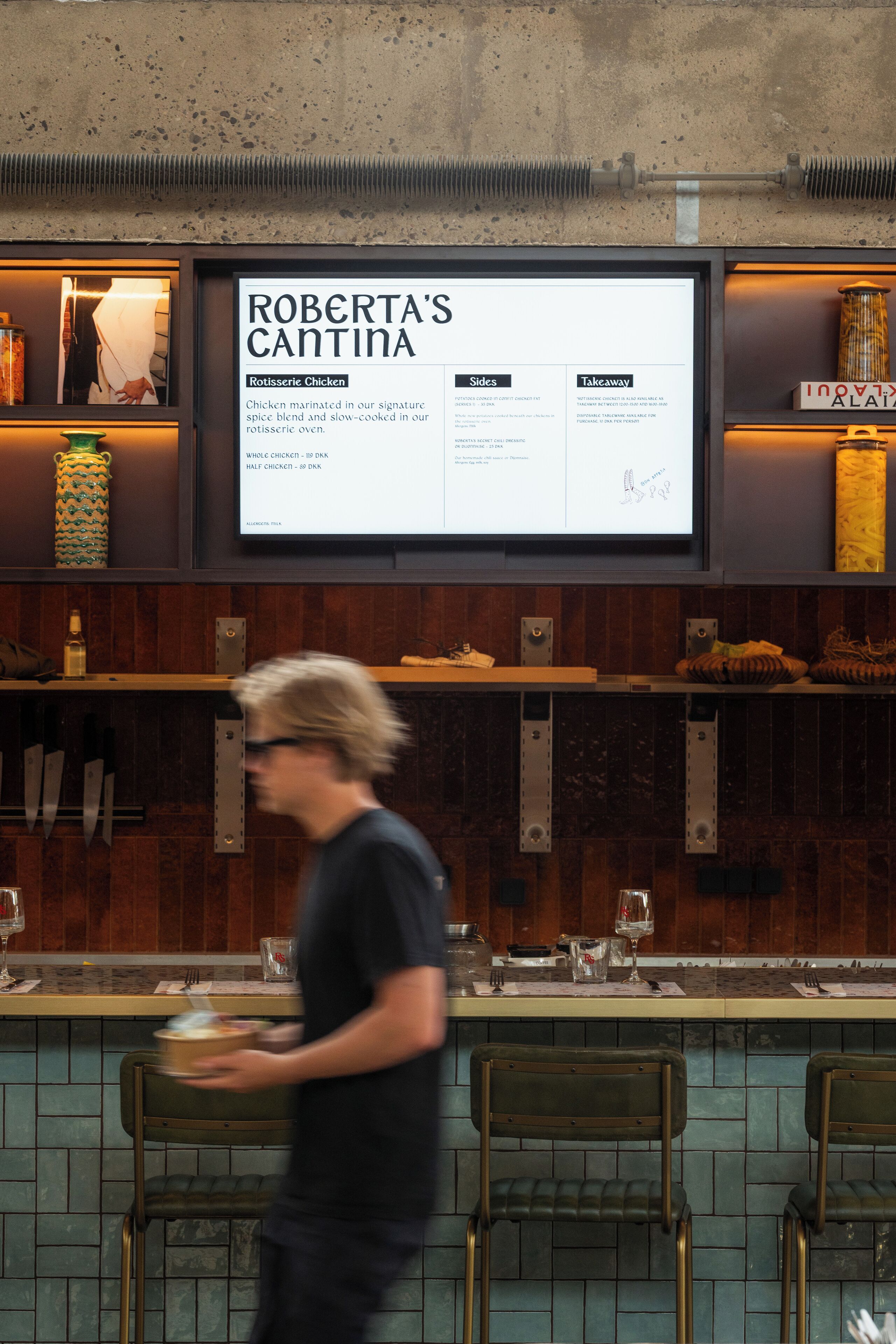 L’intérieur d’un restaurant, avec un tableau affichant le menu au-dessus du comptoir, des étagères en bois et des tabourets de bar.