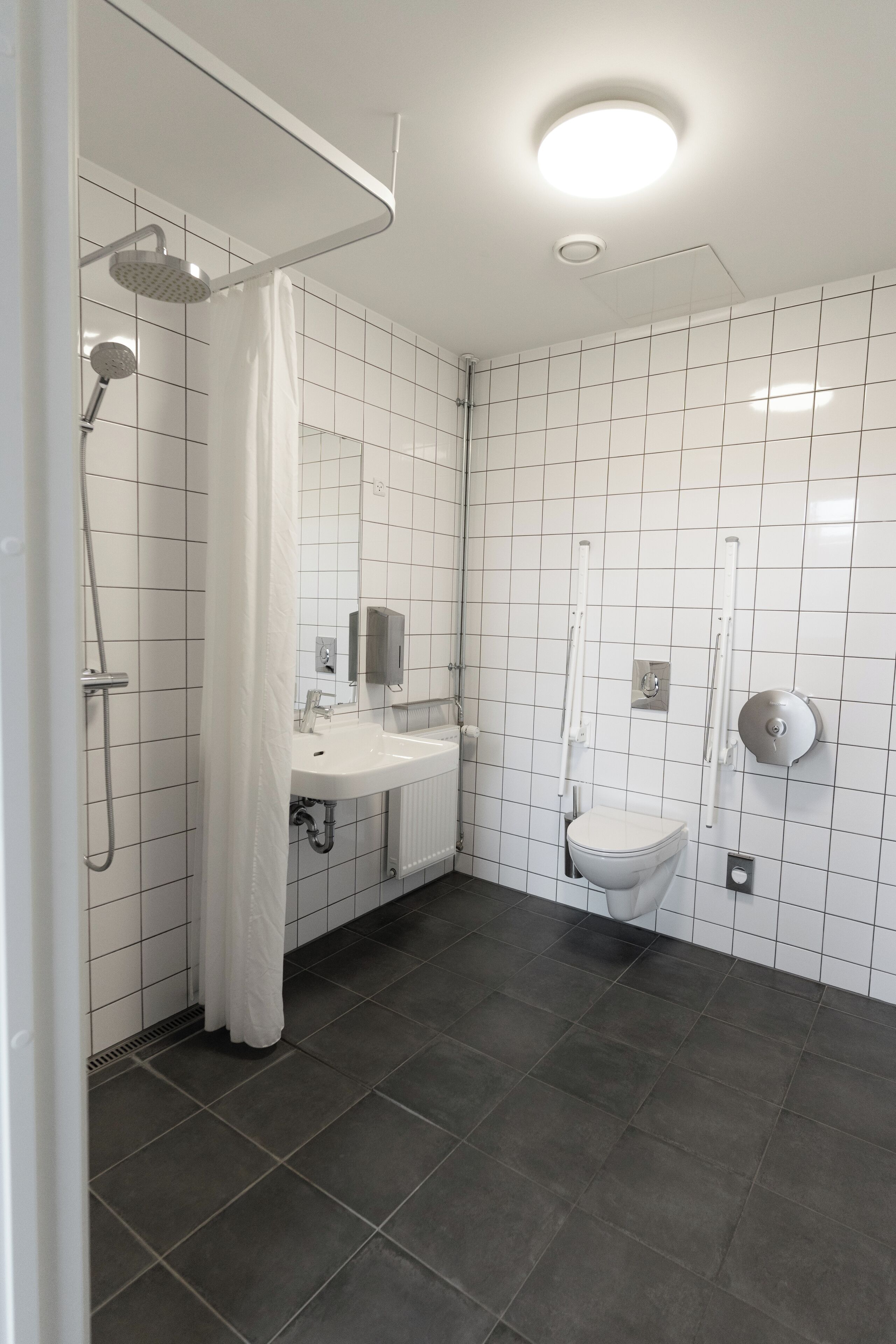 A modern bathroom with white tiled walls, a white sink, a toilet, and a shower area with a curtain.