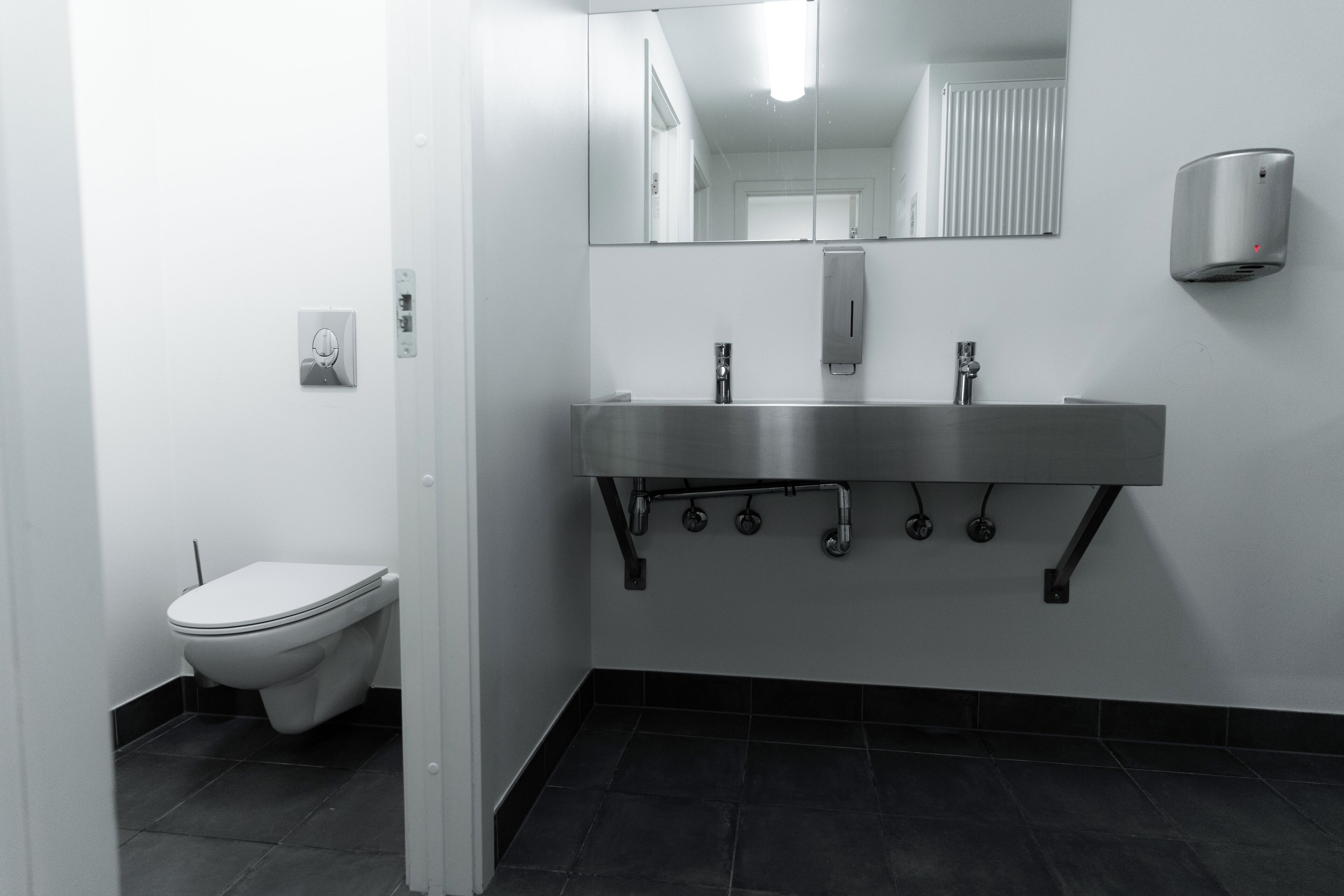 A modern bathroom with a toilet, a stainless steel sink with a mirror above it, and a hand dryer on the wall.