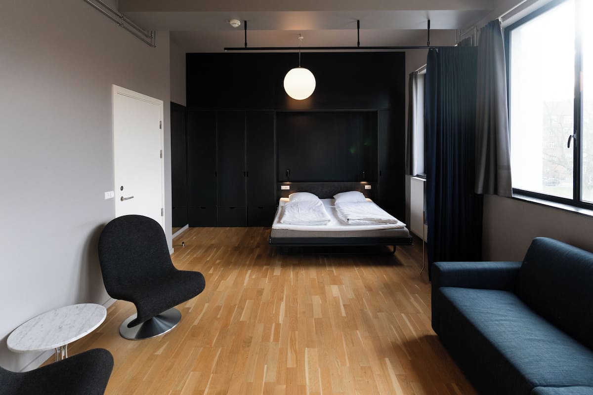 A modern hotel room with a wooden floor, a bed with white linens, a dark blue sofa, a round black chair, and a small white table.