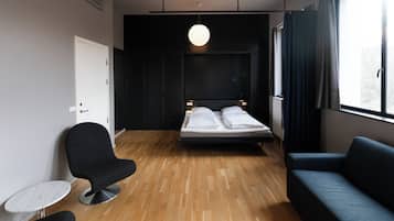 A modern hotel room with a wooden floor, a bed with white linens, a dark blue sofa, a round black chair, and a small white table.